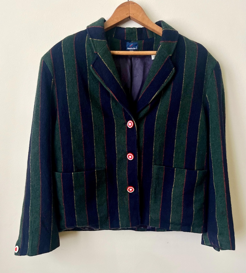 May include: A green and navy blue striped blazer with red-trimmed white buttons. The blazer has a classic collar, two front pockets, and is hanging on a wooden hanger. The lining is purple.