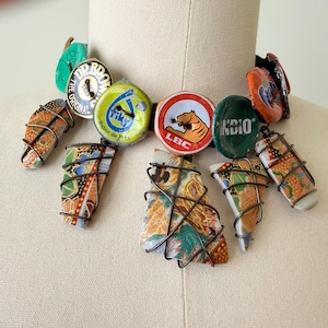 May include: A unique necklace featuring a variety of repurposed elements. The necklace includes colorful bottle caps and patterned ceramic pieces, all connected with wire. The design incorporates a mix of textures and colors, creating a bold, artistic statement.