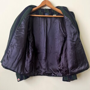 May include: A vintage blazer with a green, navy, and red plaid pattern. The jacket has a dark purple satin lining and a wooden hanger. The blazer is open, showing the interior construction and details.