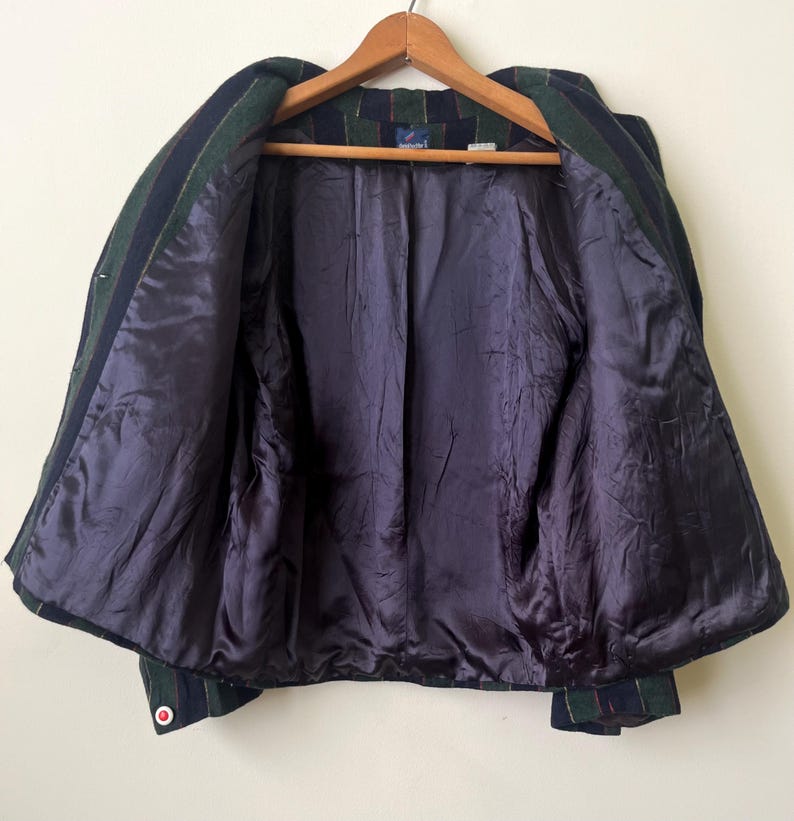 May include: A vintage blazer with a green, navy, and red plaid pattern. The jacket has a dark purple satin lining and a wooden hanger. The blazer is open, showing the interior construction and details.