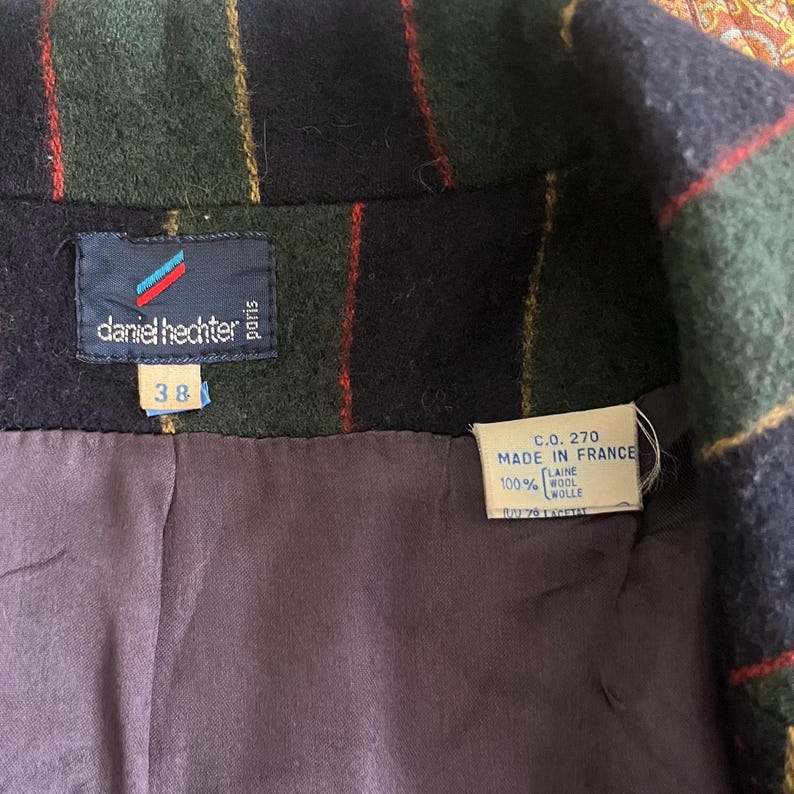May include: Close-up of a vintage wool jacket with a plaid pattern in green, navy, and red. The label reads "daniel hechter paris" and "38". The jacket is lined with a purple fabric and has a "Made in France" tag.