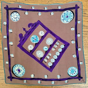 May include: A square scarf in a muted brown tone featuring a dark purple frame illustration. The design includes various white and blue patterned plates and small teacups arranged both inside the frame and along the outer border edges.
