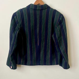 May include: A striped blazer in navy blue, green, and red. The jacket has a classic collar and button details. The vertical stripes create a tailored look. The blazer is hanging on a wooden hanger against a white background.