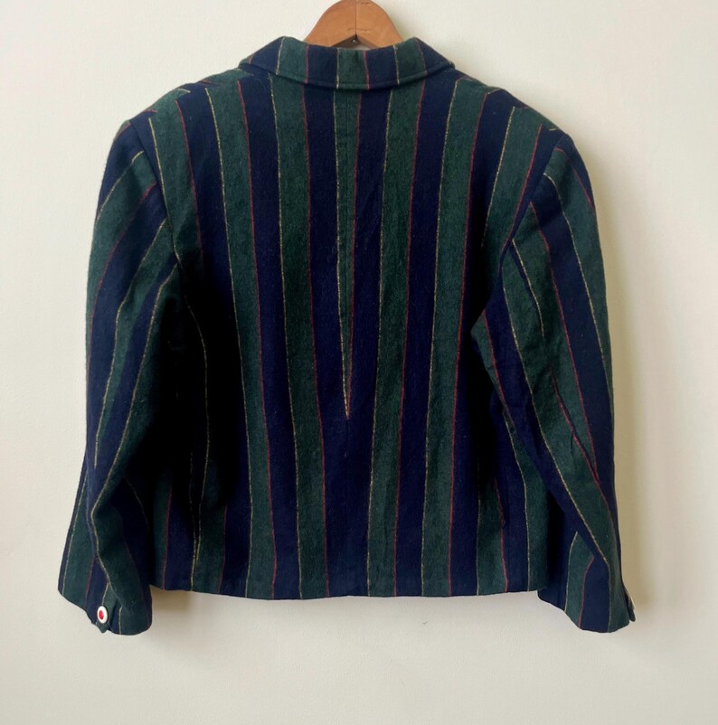 May include: A striped blazer in navy blue, green, and red. The jacket has a classic collar and button details. The vertical stripes create a tailored look. The blazer is hanging on a wooden hanger against a white background.