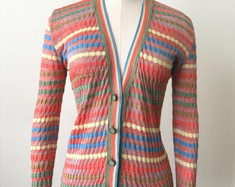 May include: A vintage, multi-colored striped cardigan sweater. The sweater features a V-neck, long sleeves, and a button-down front. The color palette includes shades of red, orange, green, blue, and yellow. The sweater is displayed on a mannequin.