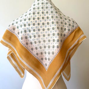May include: A square scarf featuring a white center with a repeating pattern of small blue and yellow floral motifs and dots. The scarf is framed by a wide mustard yellow border, which is accented by thin white and blue decorative line patterns.