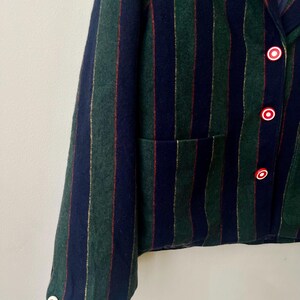 May include: A close-up of a blazer with vertical stripes in navy, green, and gold. The blazer has red and white buttons and a pocket. The fabric appears to be wool or a wool blend.