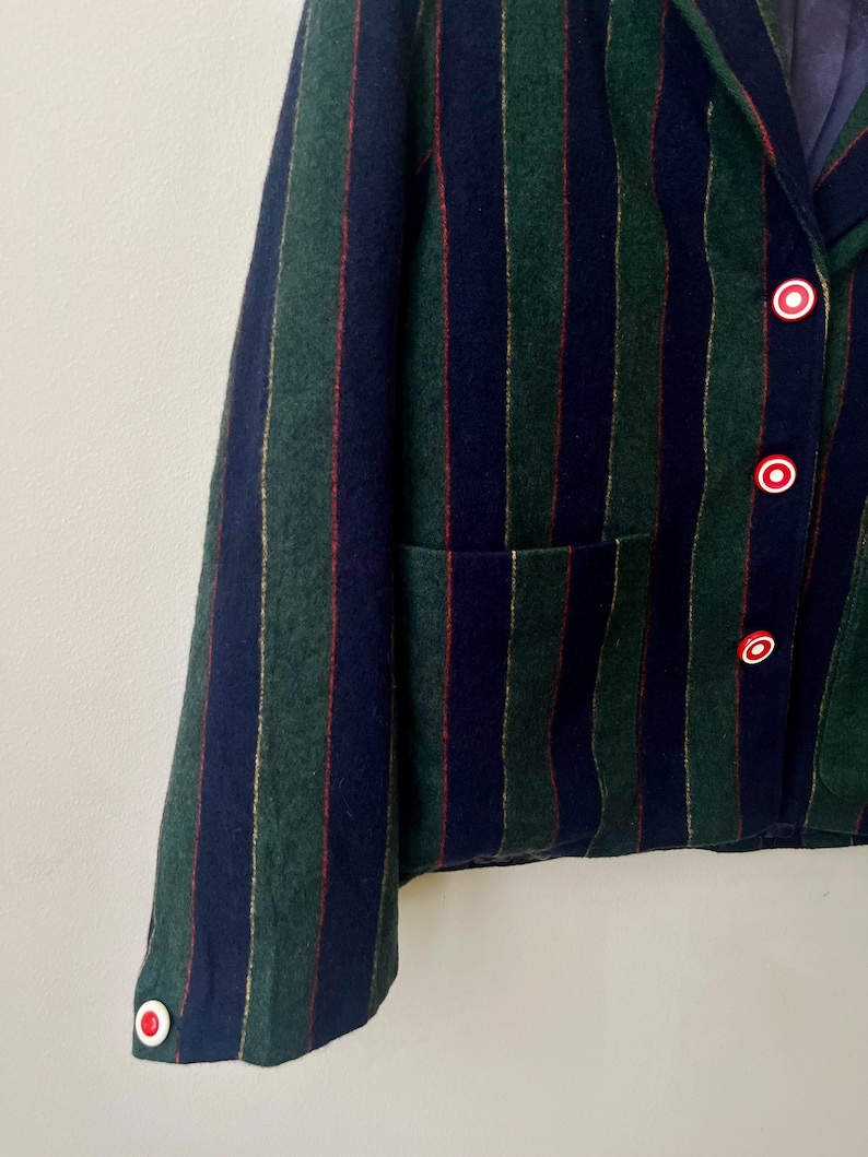 May include: A close-up of a blazer with vertical stripes in navy, green, and gold. The blazer has red and white buttons and a pocket. The fabric appears to be wool or a wool blend.