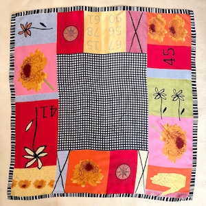May include: A colorful square scarf with a black and white striped border. The scarf features a patchwork design with sunflowers, numbers, and geometric shapes in red, pink, orange, and blue. The center is a black and white checkered pattern.