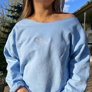 May include: A light blue off-the-shoulder sweatshirt with a white embroidered design of a shooting star with a trail of smaller stars.