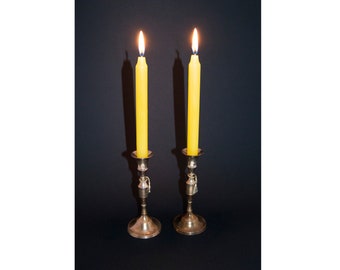 May include: Two yellow candles are lit and burning in silver candle holders on a black background.