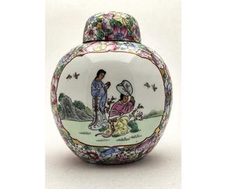 Puede incluir: Un jarrón de cerámica redondo con tapa, con una escena pintada a mano de dos figuras en un jardín. El jarrón es blanco con un borde y tapa floral, y las figuras visten trajes tradicionales.