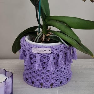Peut inclure: Une orchidée en pot avec des fleurs tachetées de violet et de blanc. La plante est dans un pot en macramé lavande fait à la main avec des glands. Le pot est sur une surface en bois.