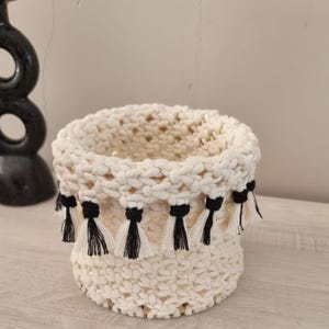 Peut inclure: Un panier en macramé de couleur crème, de forme cylindrique. Le panier présente un motif tissé et est orné de pompons noirs et crème le long du bord supérieur. Ce panier décoratif convient au rangement ou à l'exposition.