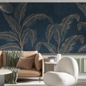 Könnte beinhalten: Ein modernes Wohnzimmer mit einer marineblauen Wand mit goldenen Bananenblattmustern. Vor der Wand befindet sich ein beiges Sofa mit einem grünen Überwurf und braunen Kissen. Ein weißer Stuhl und ein Beistelltisch vervollständigen den Raum.