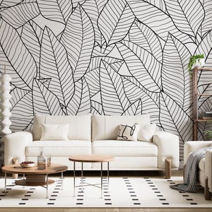 Könnte beinhalten: Ein modernes Wohnzimmer mit einem weißen Sofa, zwei runden Couchtischen aus Holz und einem gemusterten Teppich. Der Mittelpunkt ist ein großes Wandbild mit einem schwarz-weißen Blattdesign. Ein Holzregal mit Pflanzen sorgt für einen Hauch von Natur.