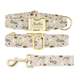 May include: A beige dog collar with a gold buckle and a personalised name tag that reads "Stella 210-658-3479". The collar features a floral pattern with bees. The matching lead is also beige with a gold clip and the same floral pattern.
