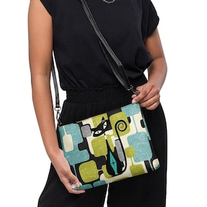 May include: A woman is wearing a black top and black pants. She is carrying a black and green crossbody bag with a cat design. The bag has a geometric pattern with blue, green, and yellow squares.
