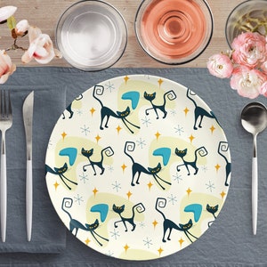 Peut inclure: Une assiette en céramique blanche avec un motif répété de chats noirs, de boomerangs bleus et d'étoiles jaunes. L'assiette est sur un set de table gris avec une fourchette et un couteau.