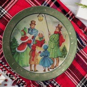 May include: A vintage Christmas plate with a green rim and a festive scene of carolers singing in the snow. The plate features a group of people dressed in winter clothing, holding songbooks and a lantern.