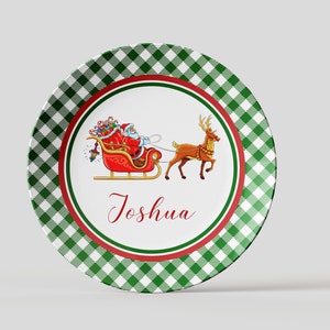 May include: A green and white checkered ceramic plate with a red border. The center of the plate features a cartoon image of Santa Claus in a red sleigh pulled by a reindeer. The text "Joshua" is written in red cursive below the image.