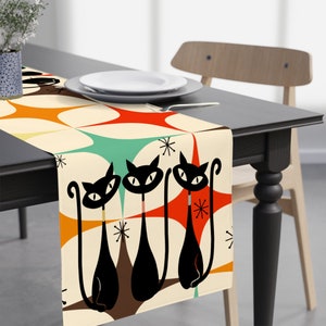 May include: A table runner with a retro design featuring three black cats with orange, green, and brown geometric shapes on a cream background.