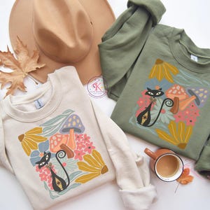 Puede incluir: Dos sudaderas, una crema y otra verde oliva, cada una con un diseño de gato negro, setas y flores. Un sombrero beige y una hoja seca sugieren un tema otoñal. Las sudaderas están dobladas.