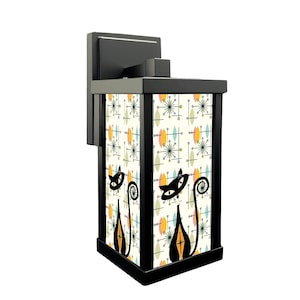 May include: Black outdoor wall sconce with a rectangular shade featuring a mid-century modern design. The shade has a cream background with a pattern of colorful starbursts and black cat silhouettes. The sconce is made of black metal.