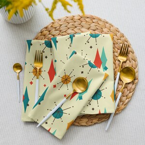 May include: A set of four yellow, blue, and red patterned napkins with a mid-century modern design. The napkins are folded and arranged on a woven straw placemat.