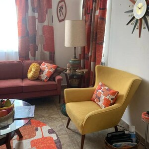 May include: A living room with a red sofa, a yellow armchair, and a mid-century modern lamp. The room has a patterned rug and curtains.