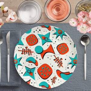 Könnte beinhalten: Ein weißer Speiseteller mit einem modernen Design aus der Mitte des Jahrhunderts mit abstrakten Formen in Tealblau und Orange. Der Teller liegt auf einem grauen Platzdeckchen mit einer weißen Gabel und einem weißen Messer.