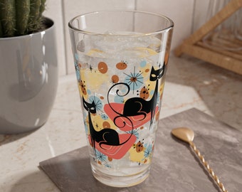 Puede incluir: Un vaso alto con un diseño retro que presenta gatos negros y formas abstractas de colores. El vaso está lleno de hielo y un líquido transparente.
