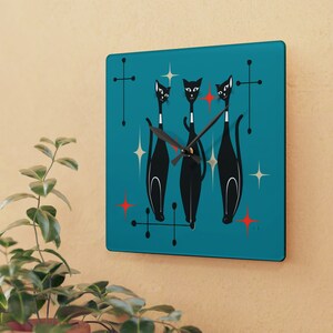 Könnte beinhalten: Eine quadratische Uhr in Türkisblau mit drei schwarzen Katzen und roten und weißen Sternen. Die Zeiger der Uhr sind schwarz.