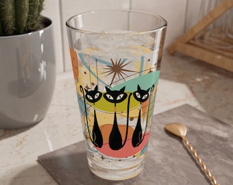May include: A tall glass with a retro design featuring three black cats with yellow eyes and tails. The glass has a colorful abstract background with a starburst pattern and is filled with ice and a clear liquid.