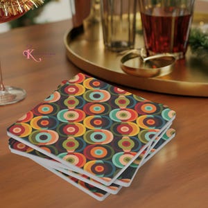 May include: A stack of square coasters featuring a repeating geometric pattern of concentric circles in shades of orange, red, teal, olive green, and dark brown. The coasters have a white base and a smooth, flat surface with rounded corners.