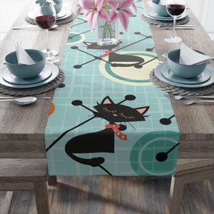 May include: A teal blue table runner with a black cat design. The cat is wearing a red and white polka dot bow tie. The background is a grid pattern with circles.