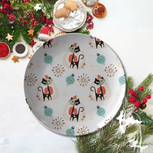 Könnte beinhalten: Ein weißer Keramikteller mit einem sich wiederholenden Muster aus schwarzen Katzen mit Weihnachtsmützen, türkisfarbenen Kugeln und Sternen. Der Teller hat ein Retro-Weihnachtsthema, ideal für Feiertagsleckereien.