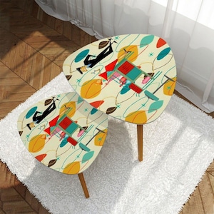 May include: Two retro-style side tables with a cream-colored surface. The tables feature a colorful mid-century modern design with geometric shapes, figures, and a light brown wooden leg. The tables are placed on a white rug.