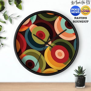 Puede incluir: Un reloj con marco negro con un patrón geométrico colorido de círculos superpuestos en tonos de rojo, naranja, amarillo, verde, azul y marrón. La esfera del reloj es negra con manecillas blancas.