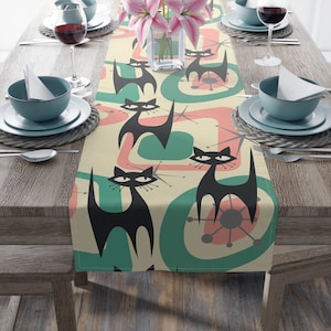 May include: A beige table runner with a retro design featuring black cats, pink and green abstract shapes, and white stars.