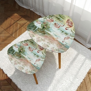 May include: Two nesting tables featuring a tropical flamingo print. The tables are made of wood and have a light brown finish. The top table is slightly larger than the bottom table.