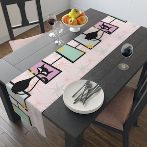 May include: A pink and green table runner with a black cat design. The runner features a repeating pattern of black cats in pink and green squares with gold stars on a pink background.