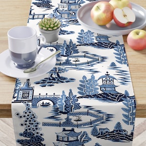 May include: Blue and white table runner with a repeating pattern of Chinese pagodas, bridges, and trees. The runner is on a wooden table with a white plate and a cup.