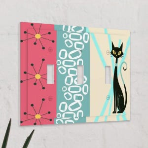 May include: Decorative light switch cover with a retro design. The cover features three sections with different patterns: pink with starbursts, teal with white shapes, and cream with a black cat illustration. The cat has yellow eyes and a teal collar.