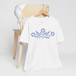 Pode incluir: Camiseta branca com um design floral azul. O gráfico apresenta três flores estilizadas com folhas e caules. Um boné de beisebol branco repousa no encosto de uma cadeira de madeira. A camisa está pendurada em um cabide de madeira.