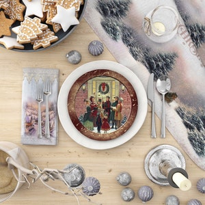 May include: A white ceramic plate with a red and gold rim features a vintage illustration of carolers singing in front of a house decorated for Christmas. The plate is on a wooden table with a white tablecloth and a silver candlestick.