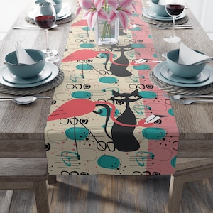 May include: A decorative table runner featuring a whimsical design with pink flamingos, black cats, and abstract shapes in teal and pink on a beige background. The runner is placed on a wooden table, with place settings and wine glasses visible.
