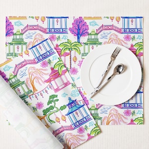 Pode incluir: Guardanapo branco com um padrão repetitivo colorido de pagodes, pontes, flores e lanternas. Um prato branco e talheres estão em cima do guardanapo.