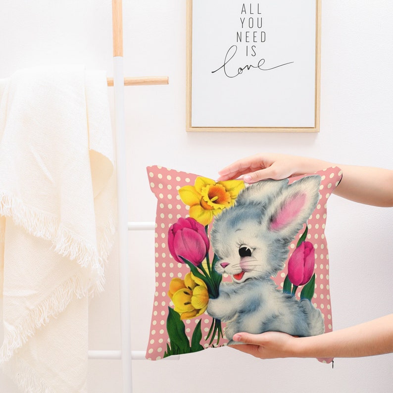 May include: A pink cushion featuring a white bunny holding a bunch of yellow daffodils and pink tulips. The background is a pink and white polka dot pattern.