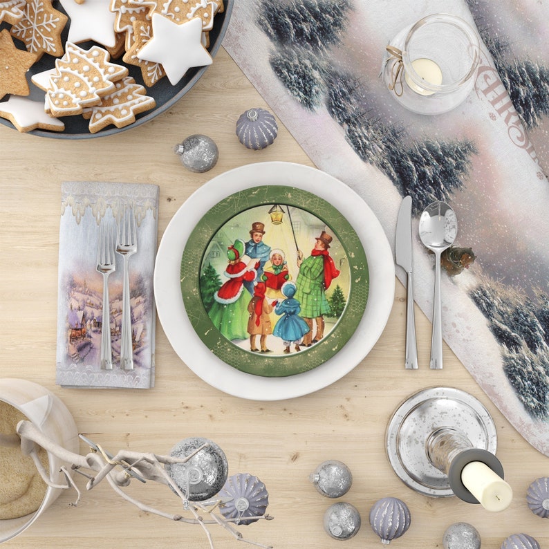 May include: A white dinner plate with a green rim and a vintage Christmas scene of people singing carols in the snow. The plate is set on a wooden table with a white napkin, silverware, and a candle holder.